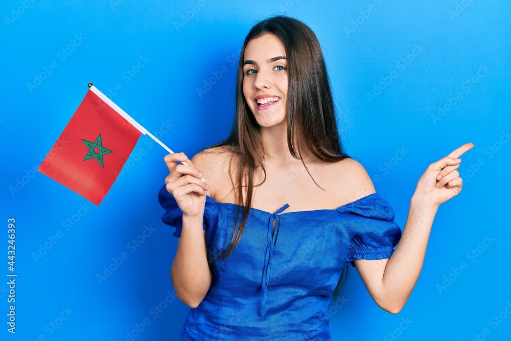 Young brunette teenager holding morocco flag smiling happy pointing with hand and finger to the side Young brunette teenager holding morocco flag smiling happy pointing with hand and finger to the side