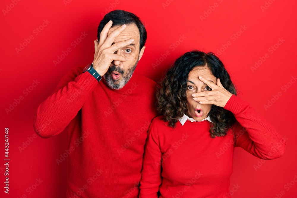Middle age couple of hispanic woman and man hugging and standing together peeking in shock covering face and eyes with hand, looking through fingers with embarrassed expression. Middle age couple of hispanic woman and man hugging and standing together peeking in shock covering face and eyes with hand, looking through fingers with embarrassed expression.