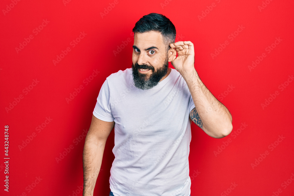 Young hispanic man wearing casual white t shirt smiling with hand over ear listening and hearing to rumor or gossip. deafness concept. Young hispanic man wearing casual white t shirt smiling with hand over ear listening and hearing to rumor or gossip. deafness concept.