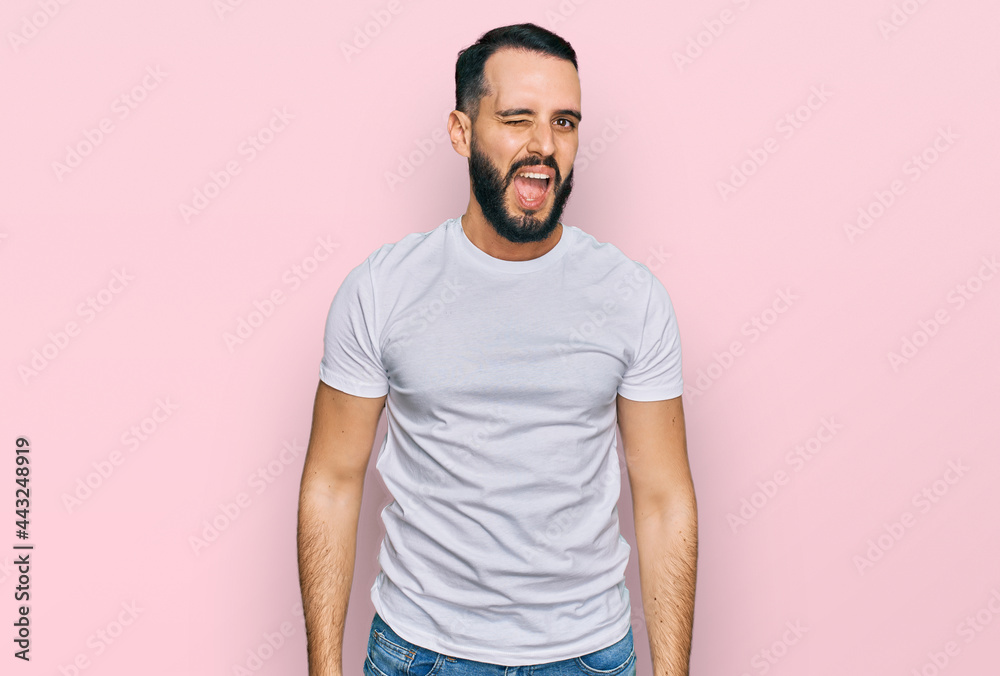 Young man with beard wearing casual white t shirt winking looking at the camera with sexy expression, cheerful and happy face. Young man with beard wearing casual white t shirt winking looking at the camera with sexy expression, cheerful and happy face.
