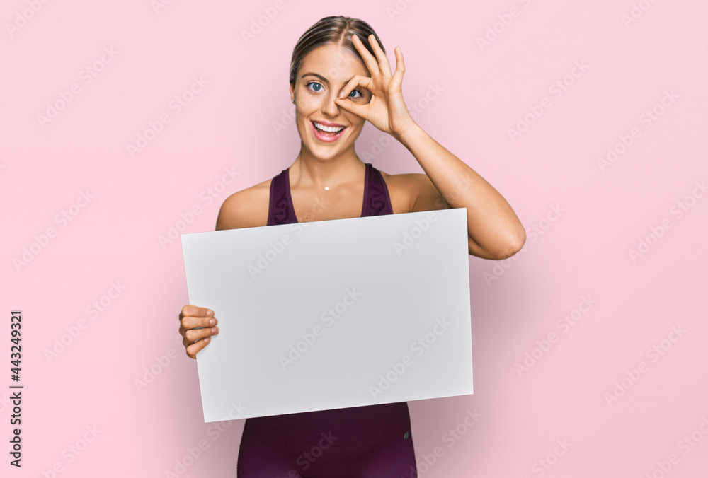 Beautiful blonde woman wearing sportswear holding white banner smiling happy doing ok sign with hand on eye looking through fingers Beautiful blonde woman wearing sportswear holding white banner smiling happy doing ok sign with hand on eye looking through fingers