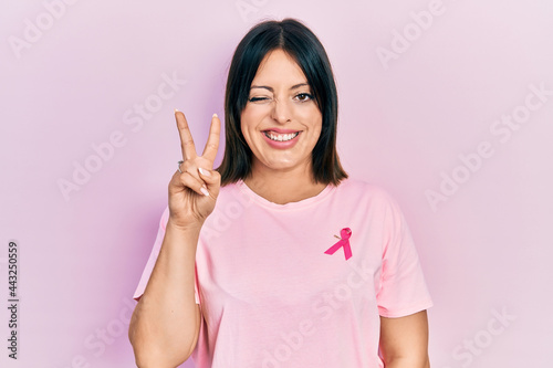 Young hispanic woman wearing pink cancer ribbon on t shirt smiling with happy face winking at the camera doing victory sign with fingers. number two.