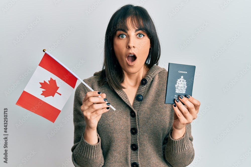Naklejka premium Young hispanic woman holding canada flag and passport afraid and shocked with surprise and amazed expression, fear and excited face.