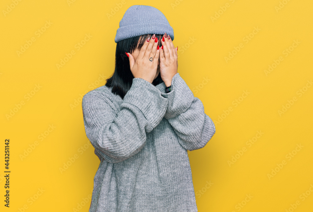 Young hispanic woman wearing cute wool cap with sad expression covering face with hands while crying. depression concept.