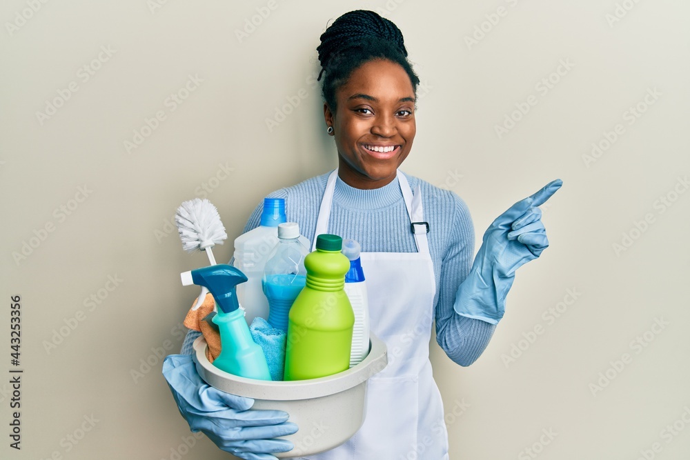 African american woman with braided hair wearing apron holding cleaning products smiling happy pointing with hand and finger to the side African american woman with braided hair wearing apron holding cleaning products smiling happy pointing with hand and finger to the side