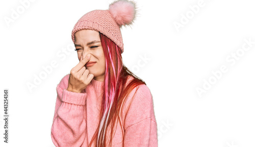 Young caucasian woman wearing casual clothes and wool cap smelling something stinky and disgusting, intolerable smell, holding breath with fingers on nose. bad smell