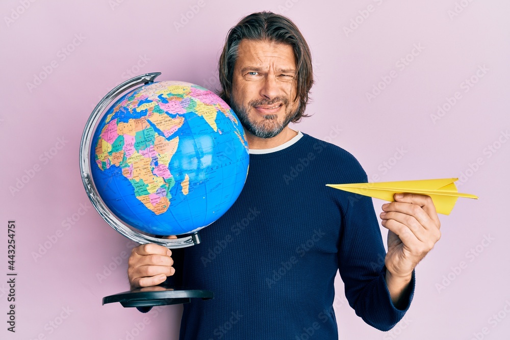 Fototapeta premium Middle age handsome man holding paper plane and world ball clueless and confused expression. doubt concept.