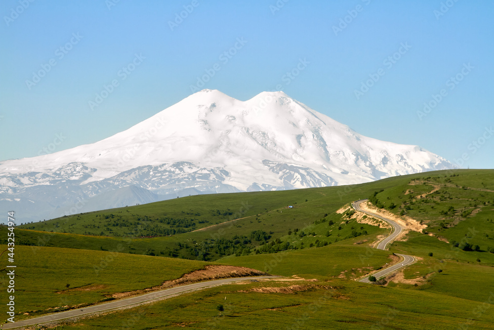 Elbrus is the highest peak in Russia and Europe. Snow mountain on the background of green fields and blue sky. Kabardino-Balkaria, view of Elbrus from Jila-Su.