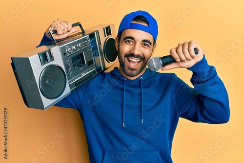 Young hispanic man holding boombox, listening to music singing with microphone smiling and laughing hard out loud because funny crazy joke.