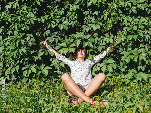 Wide smiling woman sits on grass lawn near wall all over covered with thick ivy. Natural background with green leaves and woman enjoying sunlight and warmth in urban park. Summer vibes.