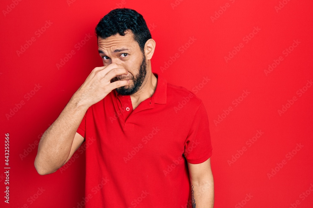 Hispanic man with beard wearing casual red t shirt smelling something stinky and disgusting, intolerable smell, holding breath with fingers on nose. bad smell Hispanic man with beard wearing casual red t shirt smelling something stinky and disgusting, intolerable smell, holding breath with fingers on nose. bad smell