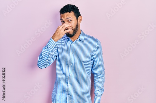 Hispanic man with beard wearing casual blue shirt smelling something stinky and disgusting, intolerable smell, holding breath with fingers on nose. bad smell © Krakenimages.com