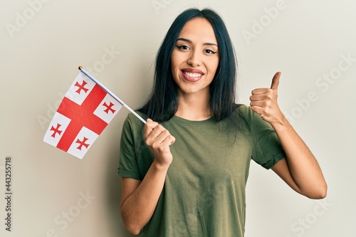 Young hispanic girl holding georgia flag smiling happy and positive, thumb up doing excellent and approval sign