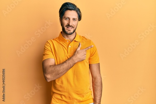Young hispanic man wearing casual yellow t shirt cheerful with a smile of face pointing with hand and finger up to the side with happy and natural expression on face
