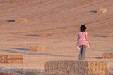 latina woman in pink t-shirt and black hair looking at a harvested field of straw bales on a bale of straw.