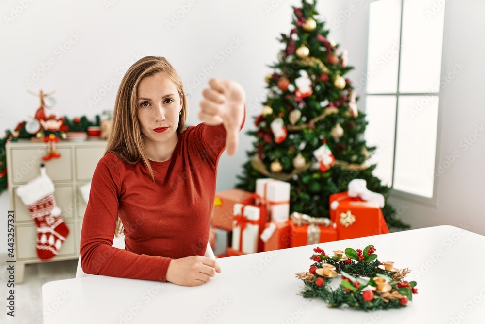 Caucasian young blonde woman sitting on the table by christmas tree looking unhappy and angry showing rejection and negative with thumbs down gesture. bad expression. Caucasian young blonde woman sitting on the table by christmas tree looking unhappy and angry showing rejection and negative with thumbs down gesture. bad expression.