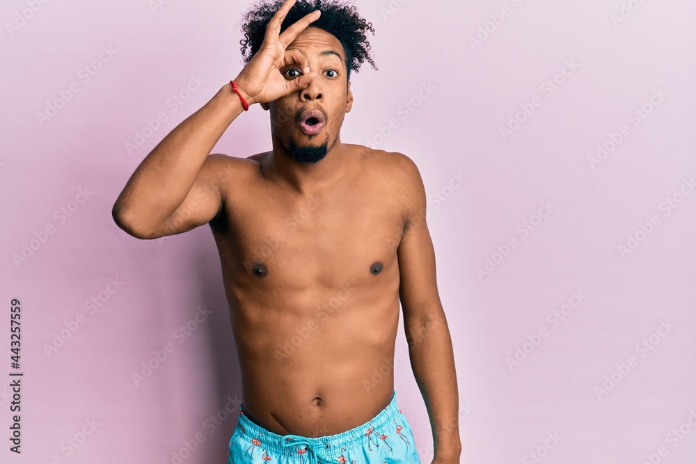 Fototapeta premium Young african american man with beard wearing swimsuit doing ok gesture shocked with surprised face, eye looking through fingers. unbelieving expression.