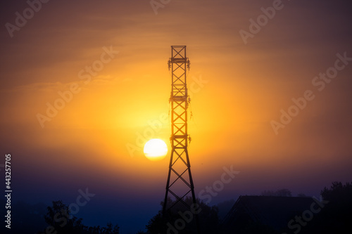 Sun rising through the mist behind a huge high voltage electricity network tower. Summertime scenery of Northern Europe.