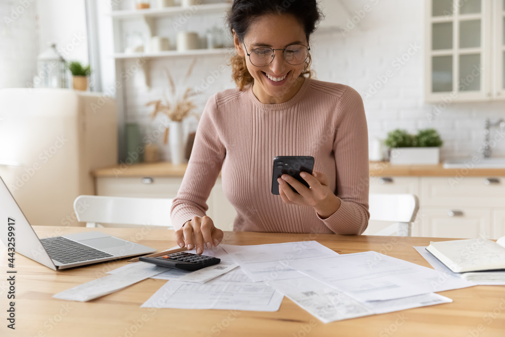Smiling young 30s woman in eyewear looking at smartphone screen, feeling satisfied with fast secure online service, paying household bills taxes or insurance, managing budget, calculating expenses. Smiling young 30s woman in eyewear looking at smartphone screen, feeling satisfied with fast secure online service, paying household bills taxes or insurance, managing budget, calculating expenses.