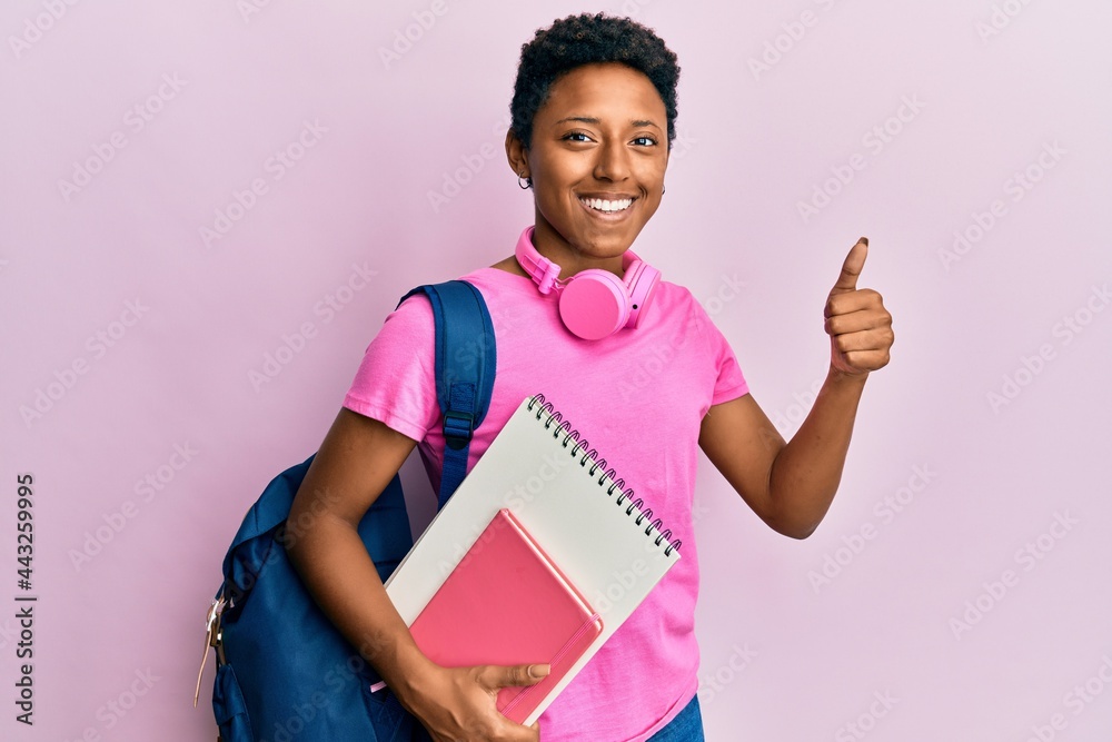Young african american girl wearing school bag and holding books smiling happy and positive, thumb up doing excellent and approval sign Young african american girl wearing school bag and holding books smiling happy and positive, thumb up doing excellent and approval sign