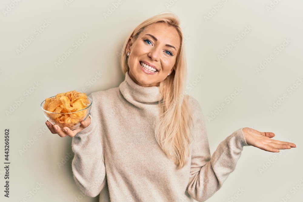 Young blonde woman holding bowl with potato chip celebrating achievement with happy smile and winner expression with raised hand Young blonde woman holding bowl with potato chip celebrating achievement with happy smile and winner expression with raised hand