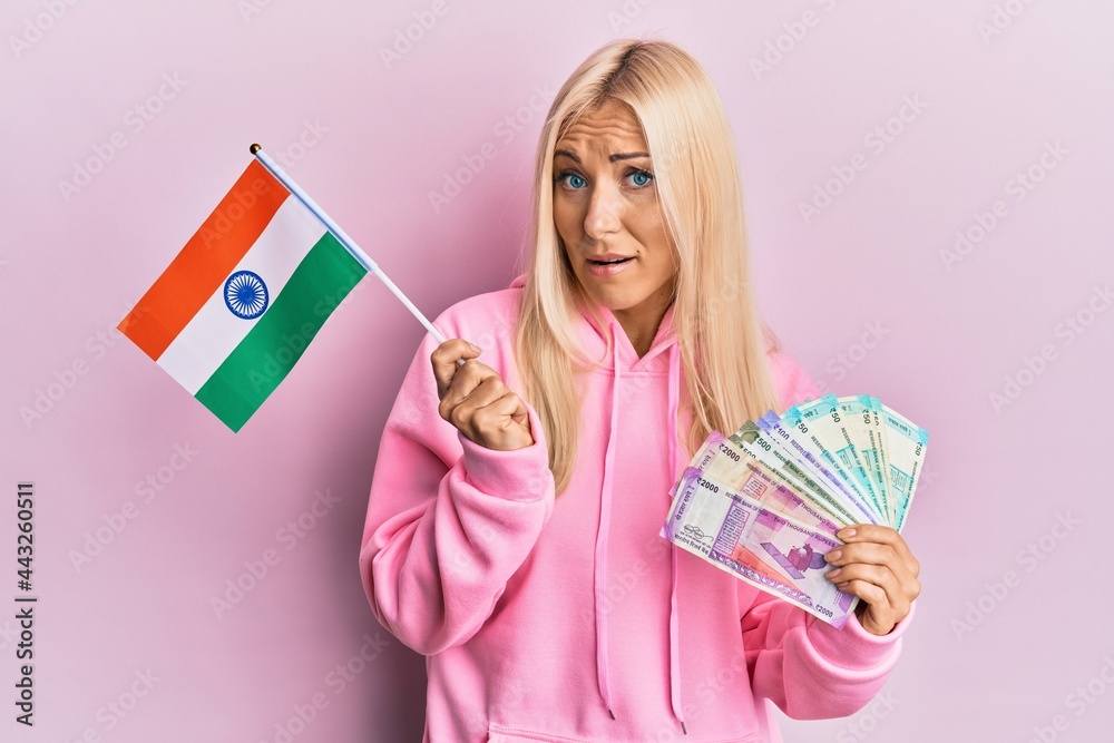 Young blonde woman holding india flag and rupee banknotes clueless and confused expression. doubt concept. Young blonde woman holding india flag and rupee banknotes clueless and confused expression. doubt concept.