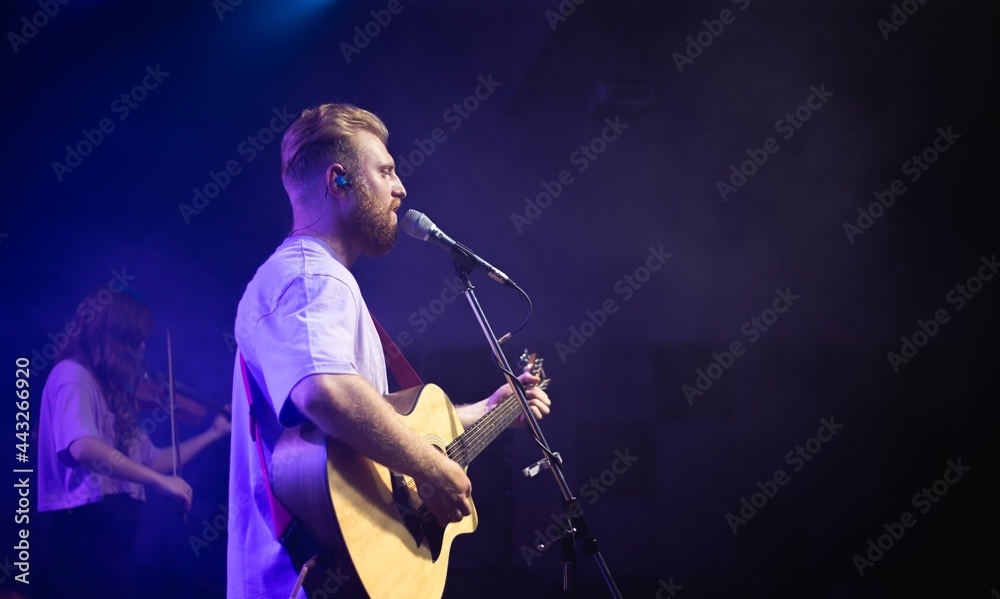 A young man with a beard holds an acoustic guitar in his hand and sings into a microphone stands on the stage