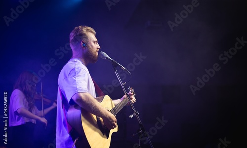 A young man with a beard holds an acoustic guitar in his hand and sings into a microphone stands on the stage