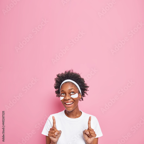 Happy dark skinned woman with curly hair applies beauty patches under eyes indicates upwards smiles gently undergoes skin care treatments dressed in whitte t shirt isolated over pink background