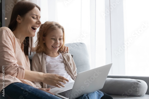 Having fun with mom. Overjoyed little schoolgirl and young female mother or elder sister watch funny comedy cartoon on pc together. Joyful mum and daughter kid laugh on cute internet memes. Copy space