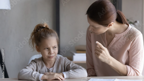 Child and parent conflict. Strict mother scolding lecturing stubborn preteen girl daughter sitting at desk reprimanding for bad behavior promise punishment. Angry mom rebuking idle kid for laziness photo