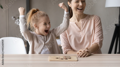 Little champion. Excited small girl daughter raise hands up scream in delight winning board game round against loving single mom. Happy elder and younger sisters play together celebrate victory of kid