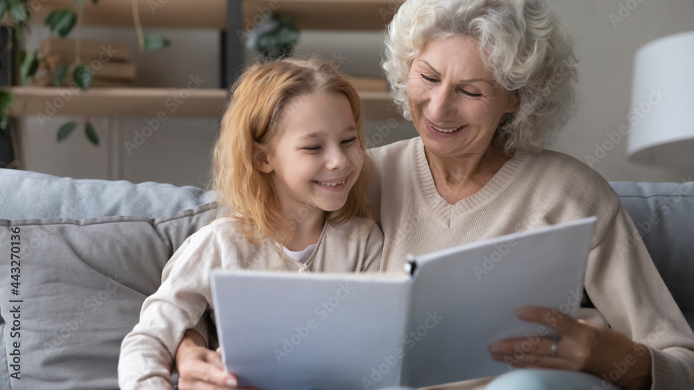 Granny storyteller. Happy smiling small granddaughter and mature grandma enjoy interesting book sitting on sofa together at leisure time. Kind elderly female baby sitter hug little girl teach to read Granny storyteller. Happy smiling small granddaughter and mature grandma enjoy interesting book sitting on sofa together at leisure time. Kind elderly female baby sitter hug little girl teach to read