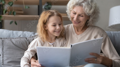 Granny storyteller. Happy smiling small granddaughter and mature grandma enjoy interesting book sitting on sofa together at leisure time. Kind elderly female baby sitter hug little girl teach to read