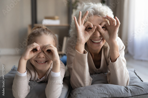 Funny goggles. Portrait of cheerful little girl grandchild lying on floor with senior granny playing having fun look at camera show binoculars glasses of fingers. Good vision and eye care for all ages photo