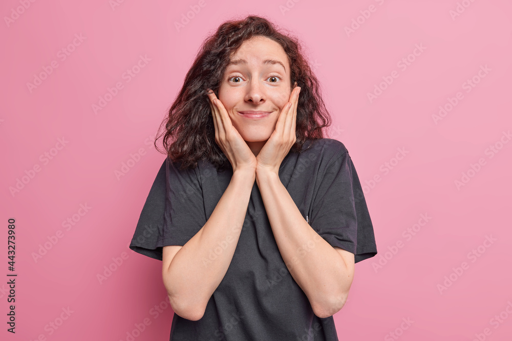 Lovely surprised young woman keeps hands on cheeks smiles pleasantly reacts on amazing news dressed in casual black t shirt isolated pink background. Happy emotions and face expressions concept Lovely surprised young woman keeps hands on cheeks smiles pleasantly reacts on amazing news dressed in casual black t shirt isolated pink background. Happy emotions and face expressions concept