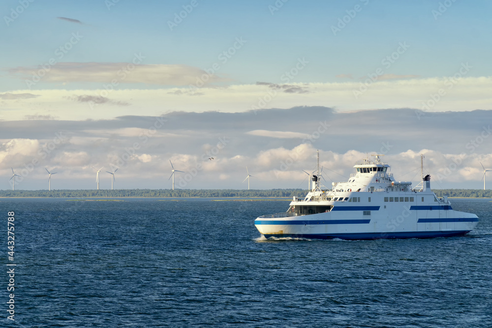 The small car ferry runs between Virtsu harbour Estonia and Saaremaa Island on Baltic Sea. Calm sea and blue sky with white clouds. In the background the wind farm on the seashore.