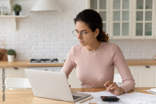 Focused millennial hispanic caucasian woman in eyeglasses calculating domestic expenses, paying bills or taxes using computer e-banking application, planning investment or checking financial data.