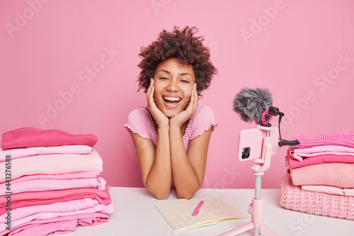 Joyful dark skined woman smiles broadly keeps hands on face records live streaming poses in front of smartphone webcam makes some notes two stacks of folded laundry near. Successful blogger. photo