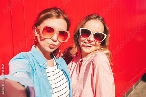 Two young beautiful smiling hipster female in trendy summer clothes.Sexy carefree women posing on street background near red colorful wall.Positive models having fun, hugging and taking Pov selfie