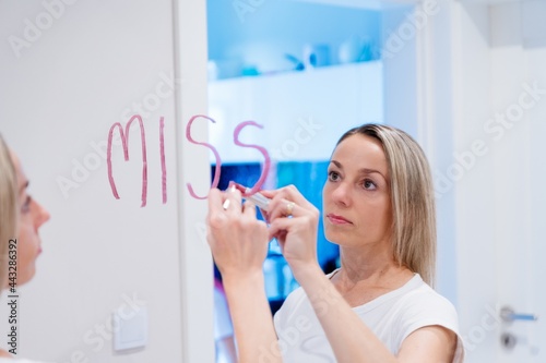 The blond woman writes the words I miss you on the mirror with a lipstick.