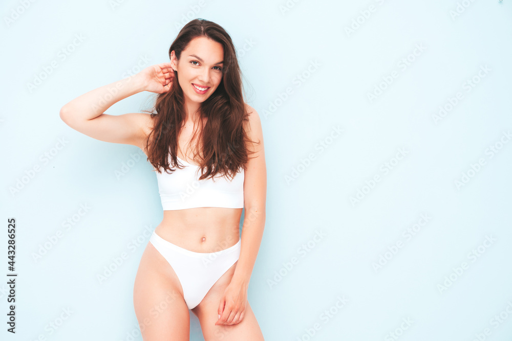 Portrait of young beautiful smiling woman in white lingerie. Sexy carefree cheerful model in underwear posing near light blue wall in studio. Positive and happy female enjoying morning