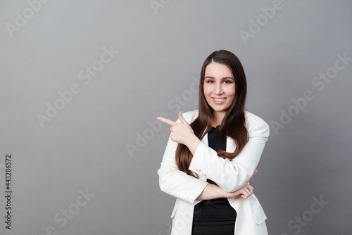 woman with long hair in a white jacket points to the side on a gray background and smiles