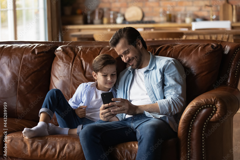 Happy kid and dad resting on cozy on couch at home, using smartphone, making video call, shopping on internet together, playing online game. Daddy and preschooler son looking at mobile phone screen Happy kid and dad resting on cozy on couch at home, using smartphone, making video call, shopping on internet together, playing online game. Daddy and preschooler son looking at mobile phone screen
