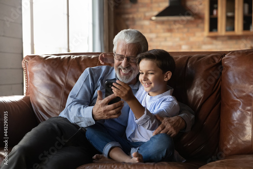 Happy preschooler boy and mature senior 70s grandpa using smartphone together, making video call, taking selfie, smiling at screen, enjoying leisure together with gadget at home, talking to family