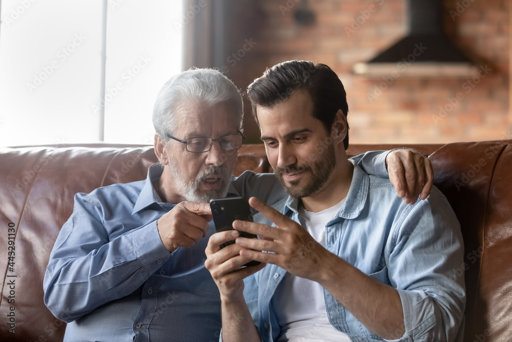 Grown son teaching elderly 70s dad to use mobile app on smartphone, showing family pictures, explaining online payment service. Two family generations men resting on couch, using cellphone together