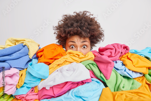 Shocked woman with curly Afro hair stares bugged eyes drowned in huge pile of colorful clothing cleans out closet selects clothes for donation or recycling white background. Housework concept
