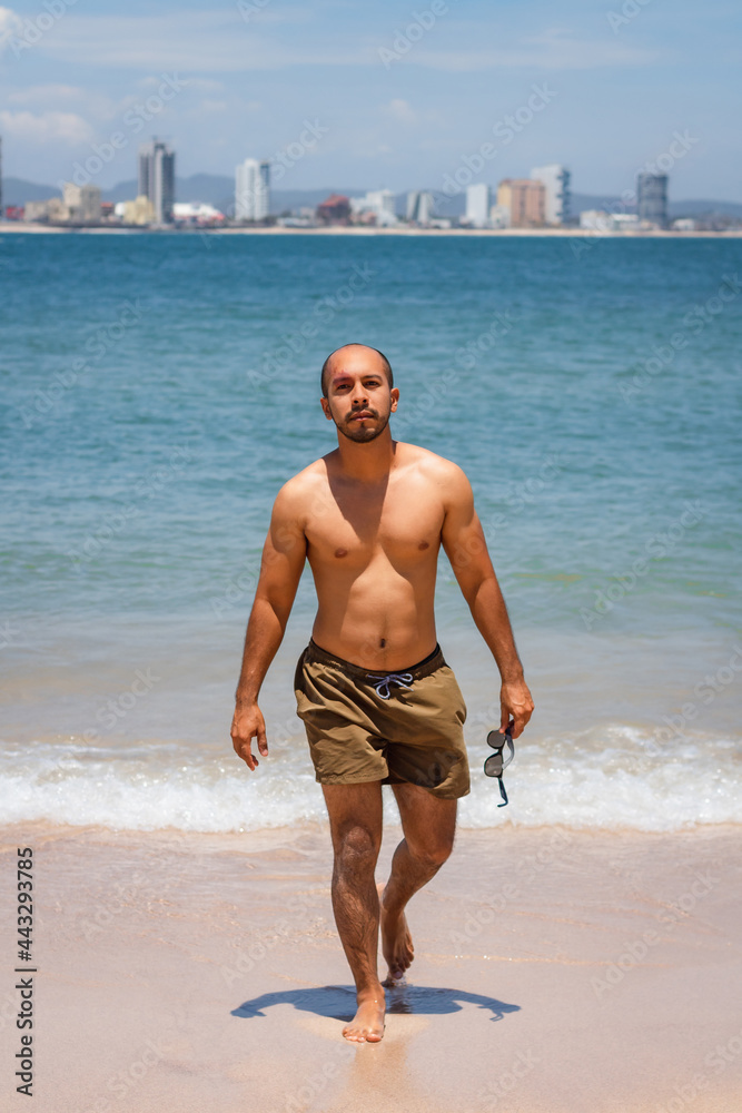 Young latin man walking looking at camera holding sunglasses at the beach