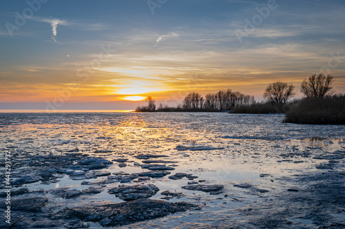Sunset on ice lake, Melting ice on the water of a lake during sunset light cloudy blue orange sky and trees on background © Lynxs Photography