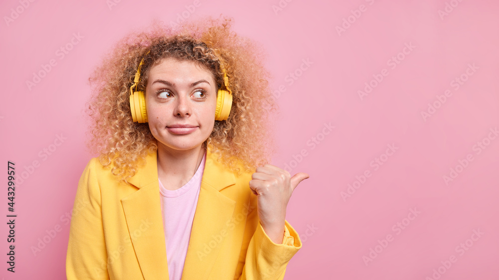 Studio shot of pretty woman with bushy curly hair points away on blank space listens music via wireless headphones dressed in formal yellow jacket shows place for your advertisement or promotion